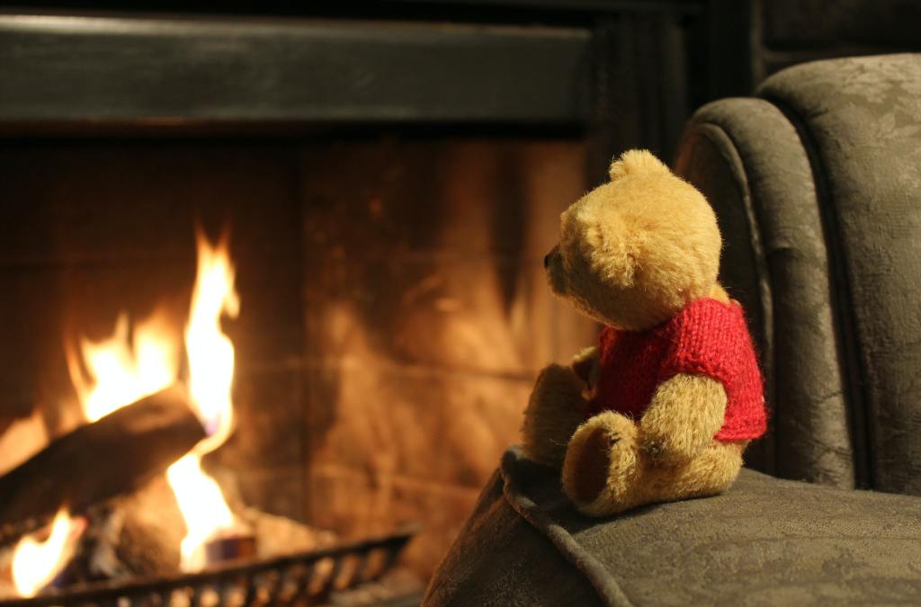 Image of small Pooh-style bear in red sweater sitting on an armchair before a fire. 