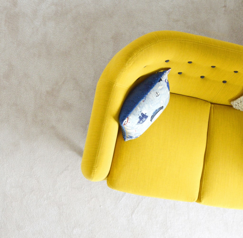 A curved yellow sofa with blue tufted buttons and a blue sailboat-themed throw pillow, shown at an angle from above.
