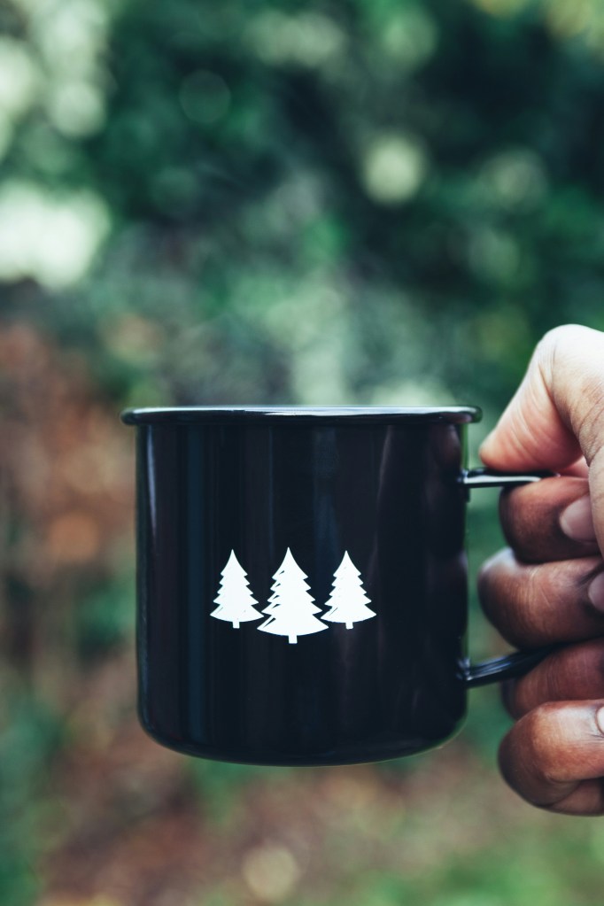 A hand holds a black enamel mug with three white evergreen trees, against a blurred background of trees.