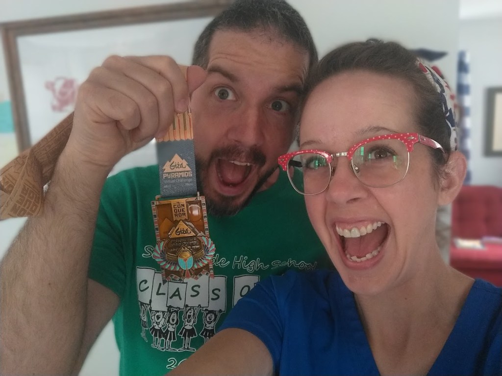 The author and her husband with exaggerated excited expressions, holding a Conquerors Giza challenge metal with turquoise scarab and pyramids.