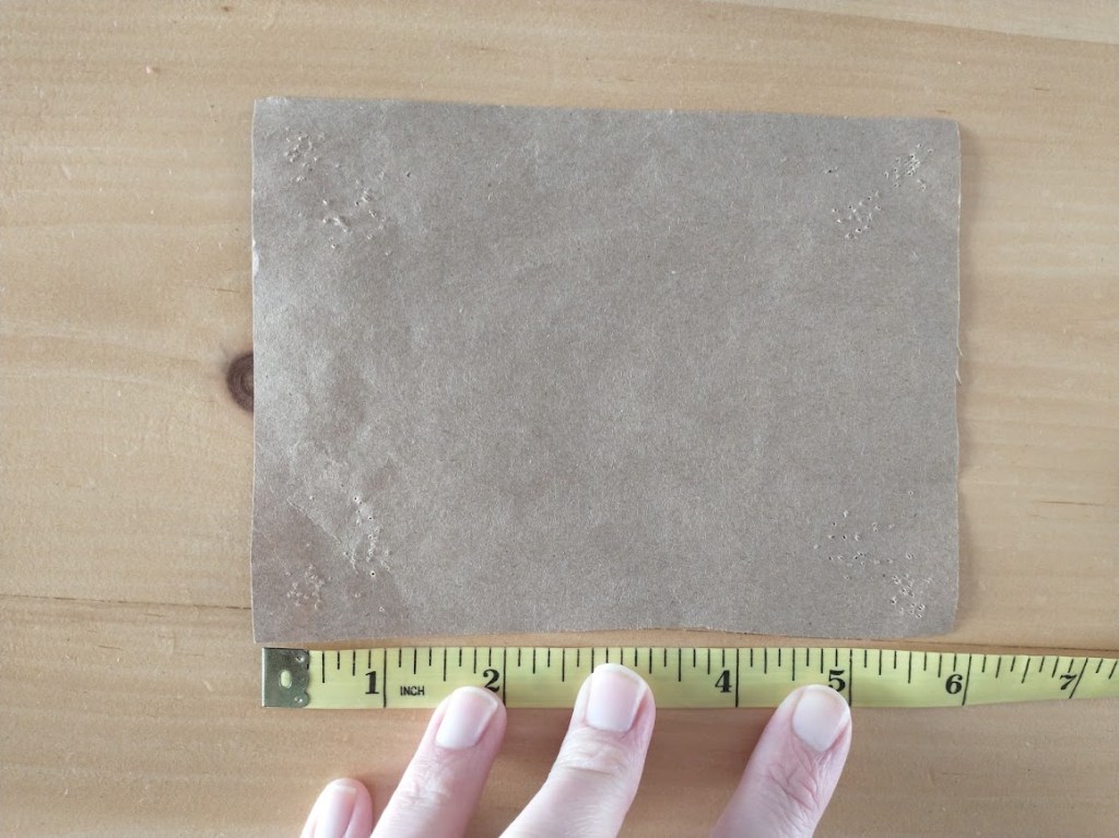 A rectangular piece of brown kraft paper, with a hand holding a measuring tape to the bottom edge, measuring 6 inches. 