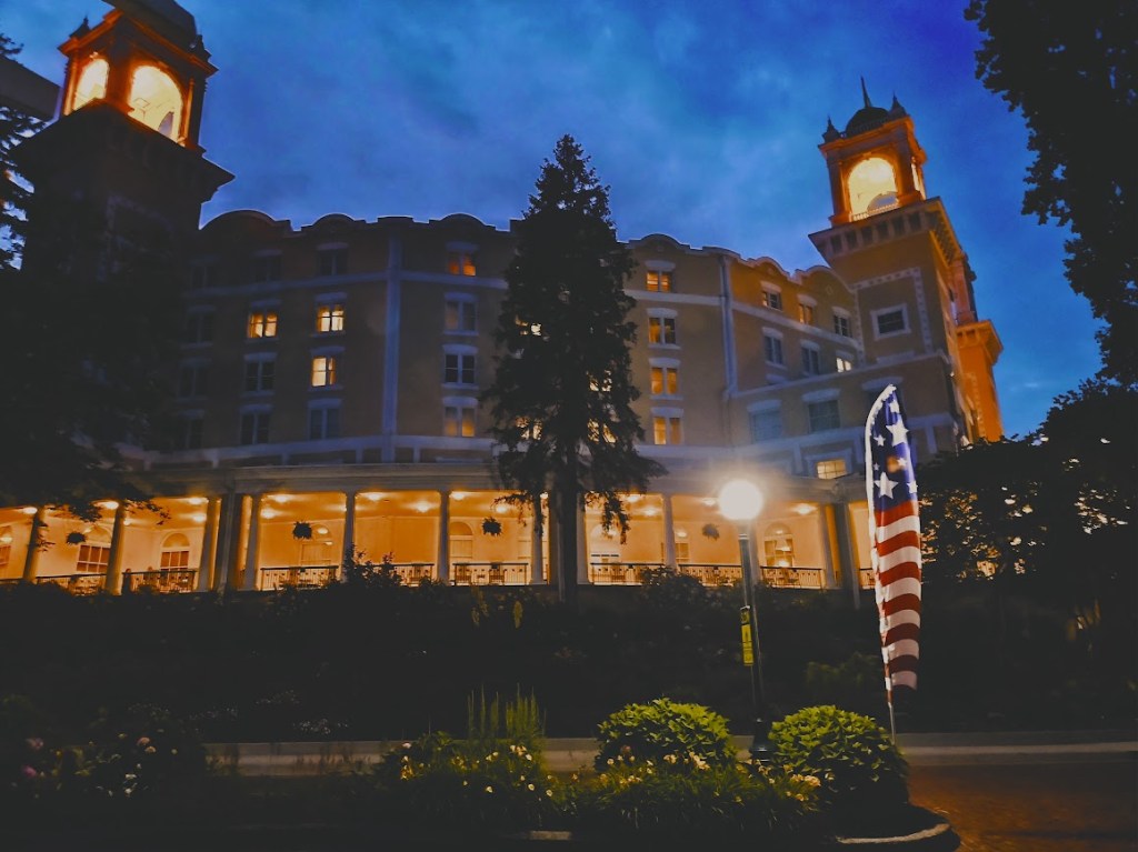 The exterior of the circular hotel, mustard-colored, with two towers at either end of the photo and large wrap-around porch, lit up against a night sky.
