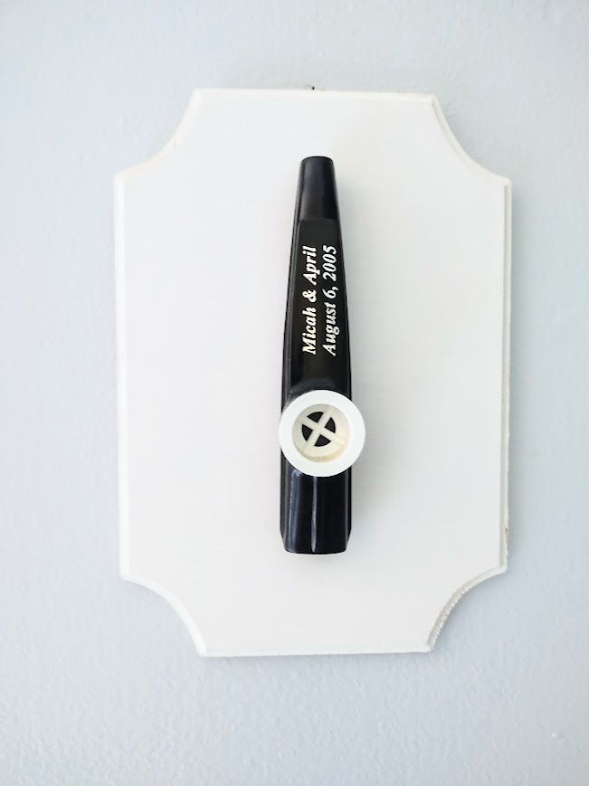 A black and white kazoo, inscribed with the author's name and wedding date, mounted on a white beveled board, hung on a pale blue-grey wall.