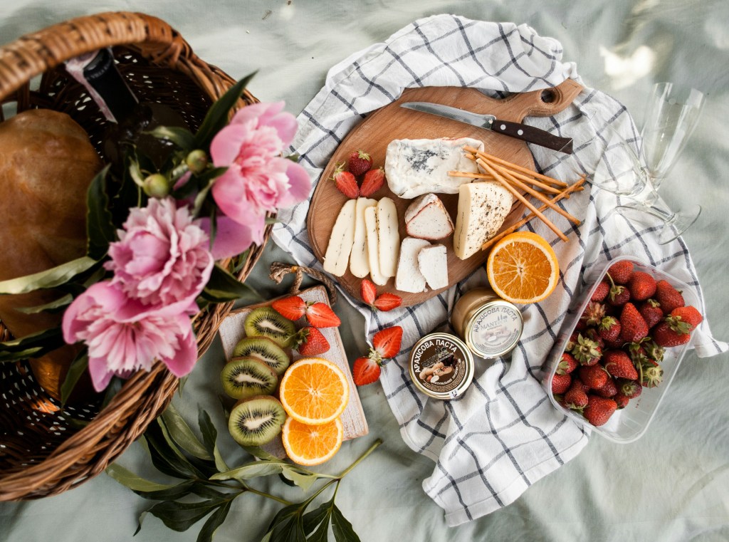 A wicker picnic basket and white blanket, spread with an assortment of fruits, cheeses, jam jars, and flowers. 