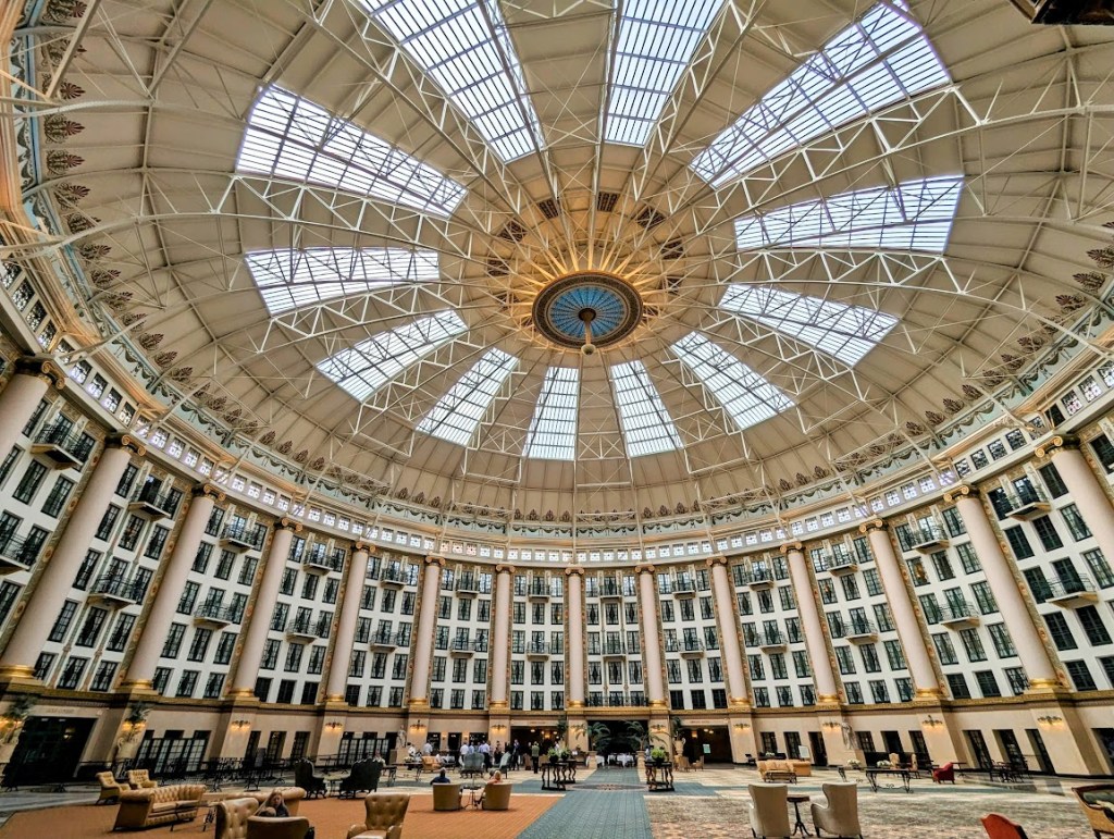 An enormous round atrium, with domed ceiling with glass sections, a Tiffany chandelier, and rows of paned windows and balcony, overlooking a large common space dotted with chairs and couches.