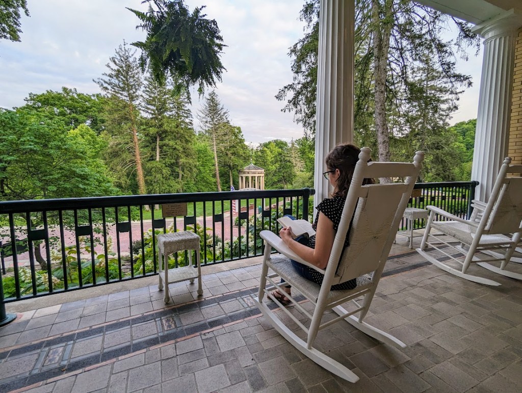 The author sits in a white rocking chair, facing outward to a garden with Greek-inspired springhouse, reading a book.