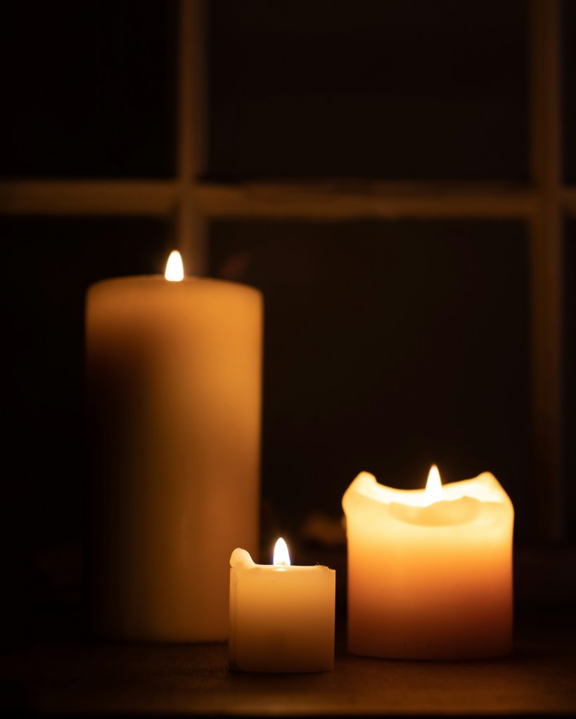 Three pillar candles burn in front of a darkened window.