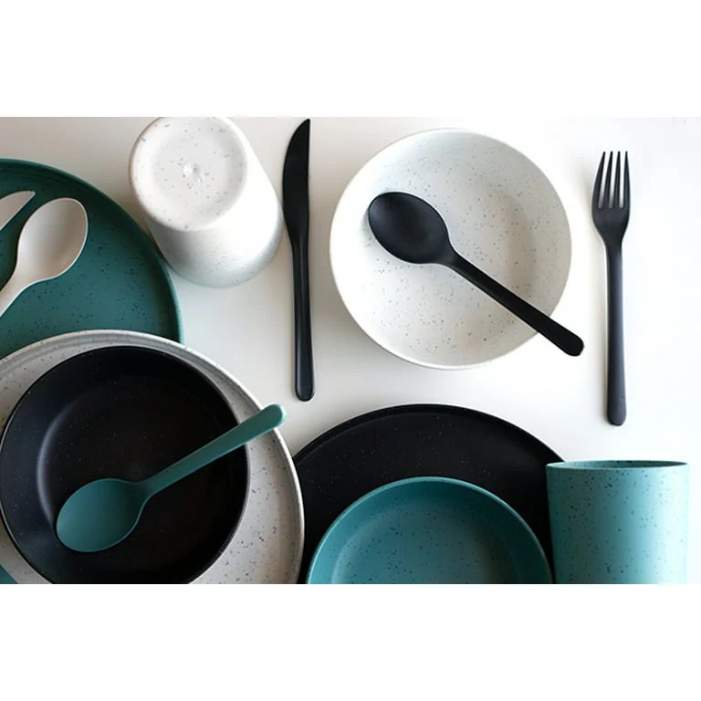 A collection of speckled matte plastic plates, bowls, cups, and cutlery, in various shades of beige, teal, and deep green are scattered on a white surface.