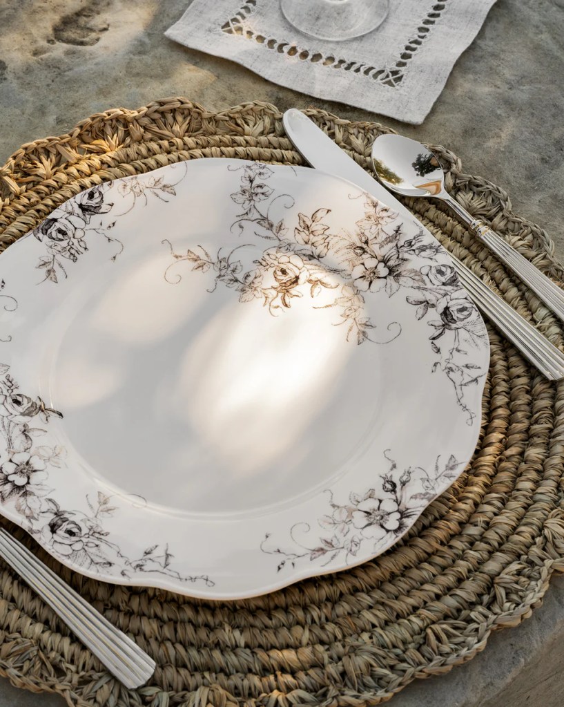A white scalloped melamine plate with sketched floral border design is shown on a woven charger with striped silver cutlery on either side and a beige linen napkin. Dappled sunlight suggests the image is taken outdoors.