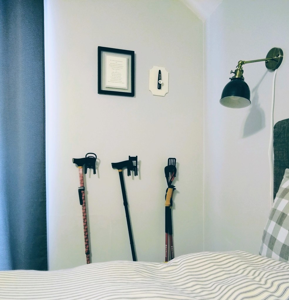 A light gray wall with three black chair-shaped hooks, with two canes and a pair of walking sticks. The edge of a bed with mattress ticking duvet, a turquoise wall lamp, and two small pieces of artwork are around them. 