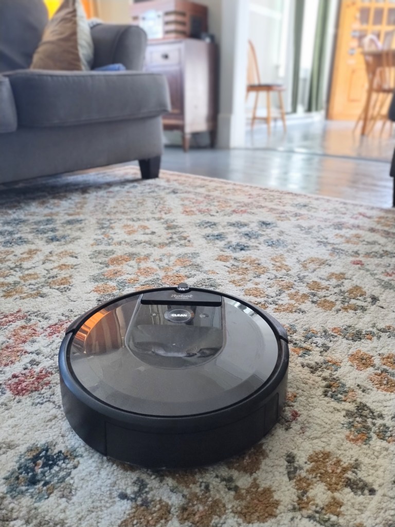 A robotic vacuum crossing a floral carpet, with a grey couch, blue wood floors, and pine dining furniture visible in the background.