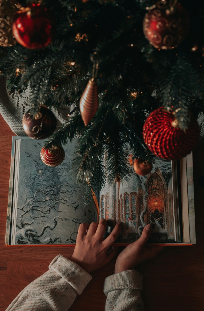 A Caucasian woman's hands hold an illustrated book (with image of a snowy scene and church-like building) under a Christmas tree with red and gold ornaments. Photo credit: Zoe, Unsplash